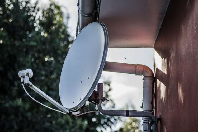A closeup shot of a white satellite dish on fixed on a brown wall with trees on the background